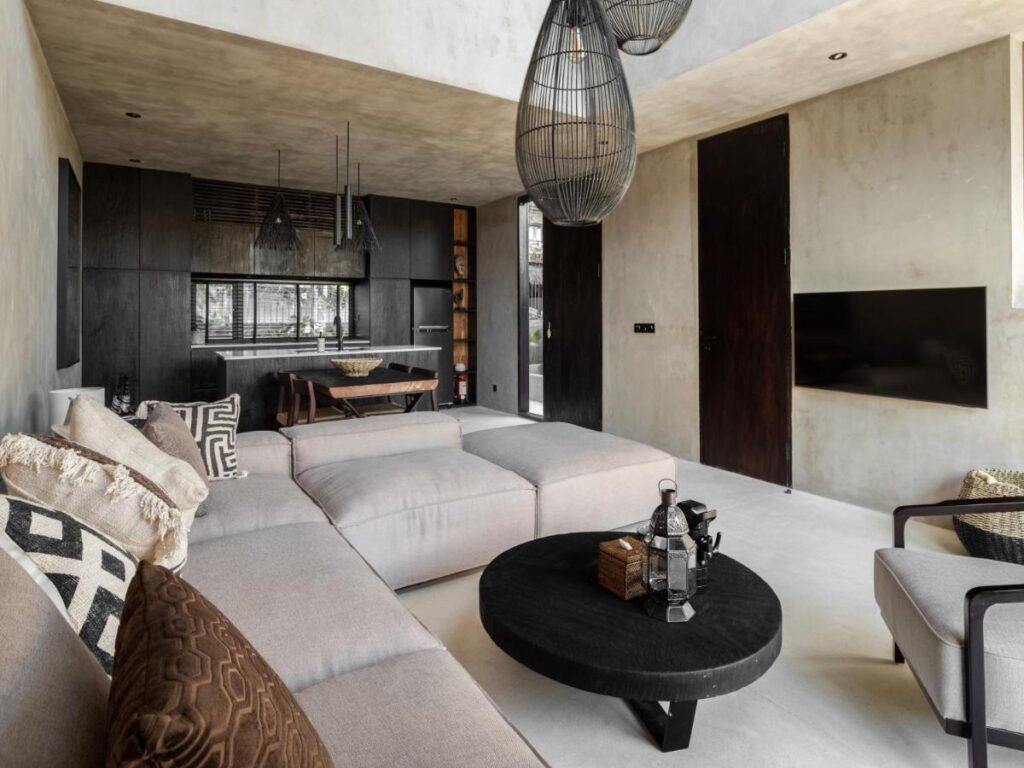 14 UNIQUE Boutique Hotels in Canggu, Bali 52 A modern living room with a large beige sectional sofa, black round coffee table, neutral-toned walls, and open kitchen—reminiscent of boutique hotels in Canggu. Pendant lights hang above, with a wall-mounted TV and armchair completing the look.