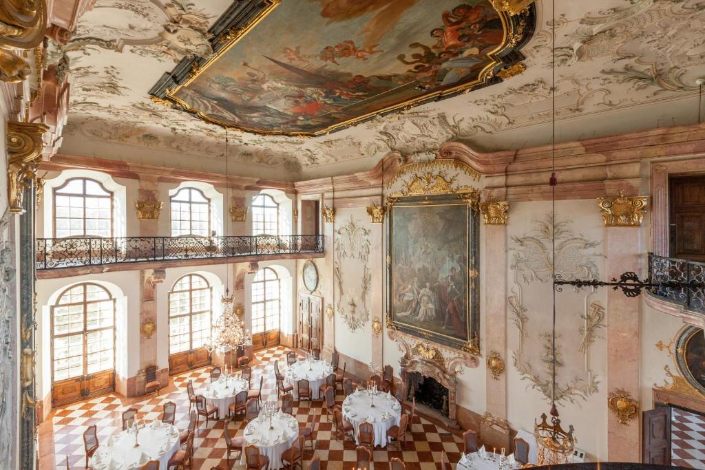 Unique Boutique Hotels in Salzburg 11 An ornate ballroom in the style of the Boutique Hotels in Salzburg, with high decorated ceilings, large arched windows, chandeliers, intricate wall art, and round dining tables atop a checkered floor. Balconies grace the upper walls.