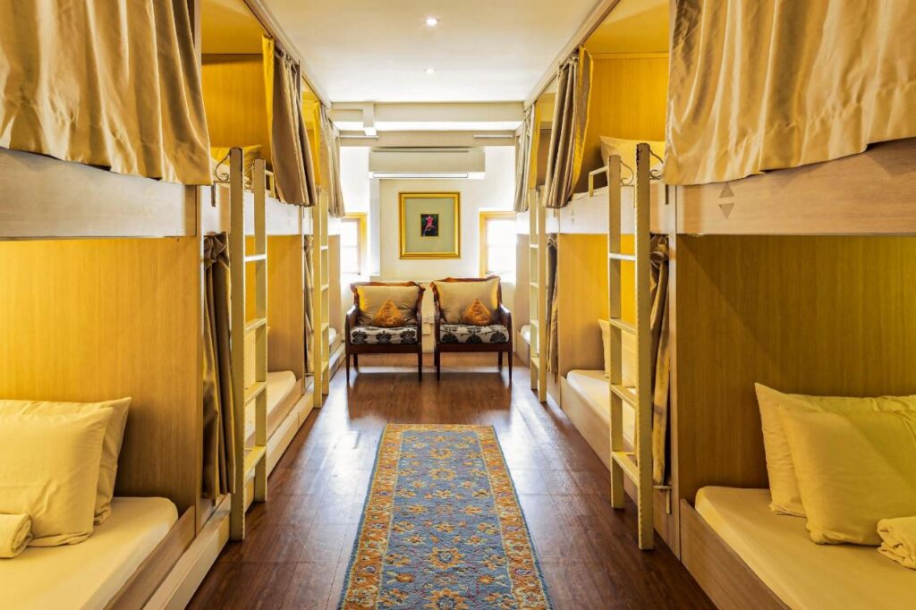 13 Most Unique Hotels in Singapore for an Incredible Holiday 46 A cozy dorm room in one of the unique hotels in Singapore, featuring four bunk beds, yellow walls, privacy curtains, a patterned blue rug at the center, and a small seating area with two chairs and cushions beneath a framed picture.
