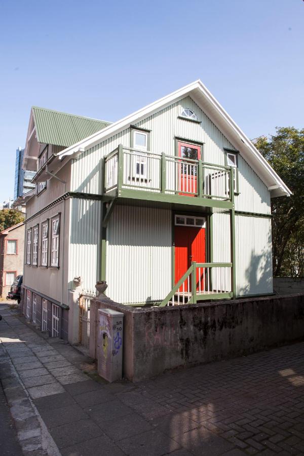 Where to Stay in Reykjavík: Top Guide 21 A two-story house with a green metal roof, green trim, a red front door, and an upper balcony is an inviting option if you’re searching for where to stay in Reykjavik. Light siding and a raised foundation sit beside a paved walkway with trees in the background.