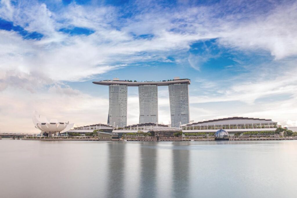 13 Most Unique Hotels in Singapore for an Incredible Holiday 7 The image shows Marina Bay Sands resort, one of the most unique hotels in Singapore, featuring three tall towers connected by a boat-shaped rooftop, with the ArtScience Museum and waterfront visible under a partly cloudy sky.