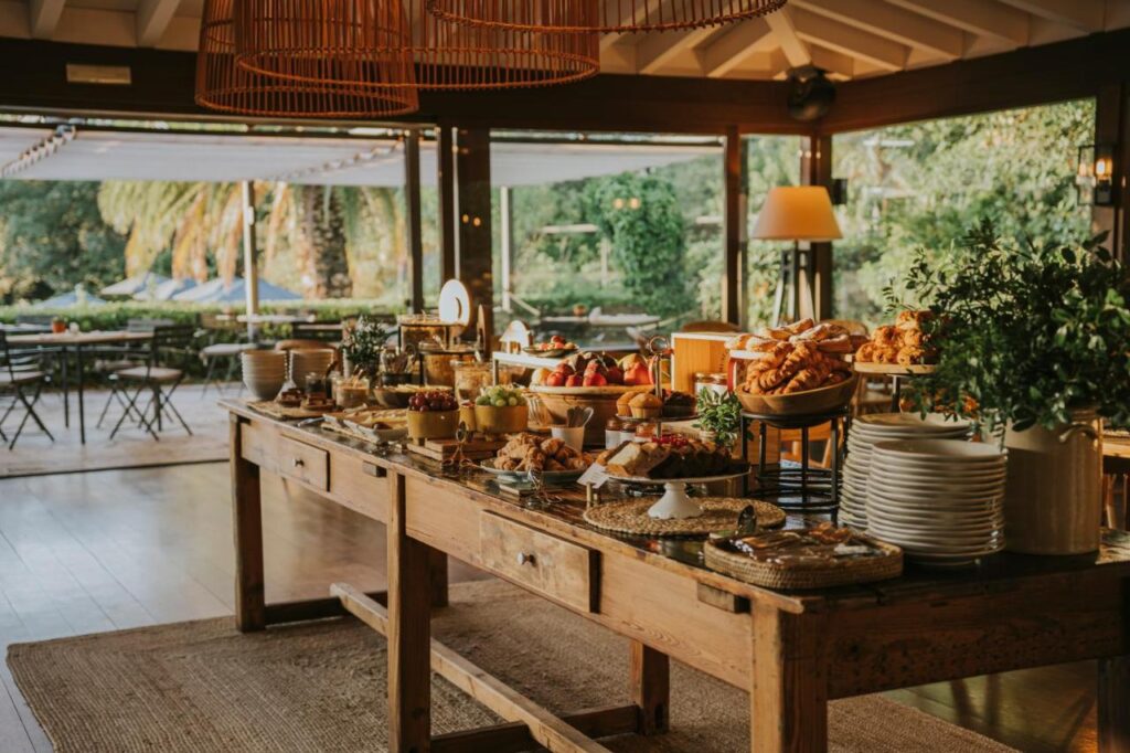 A rustic wooden table, like those found in boutique hotels in Costa Brava, is set with breakfast foods—pastries, fruit, and plates—indoors near large windows overlooking a patio and lush greenery.