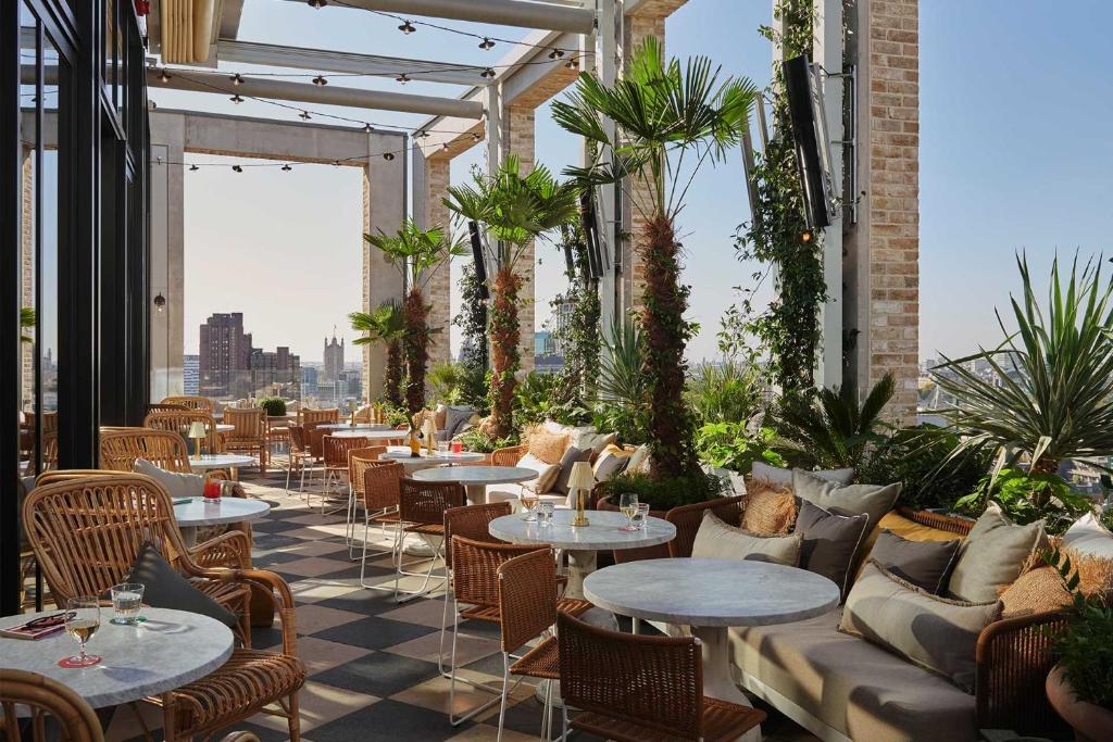 15 Best Boutique Hotels in London, UK 60 A stylish rooftop terrace at one of the boutique hotels in London features round tables, wicker chairs, and cushioned sofas surrounded by palm trees and lush plants, all overlooking the city skyline on a sunny day.
