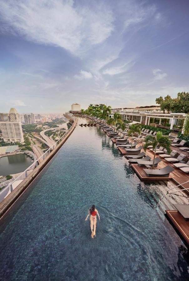 13 Most Unique Hotels in Singapore for an Incredible Holiday 5 A woman swims alone in a rooftop infinity pool lined with lounge chairs, overlooking a city skyline—one of the unique hotels in Singapore—set against distant skyscrapers and a partly cloudy sky.
