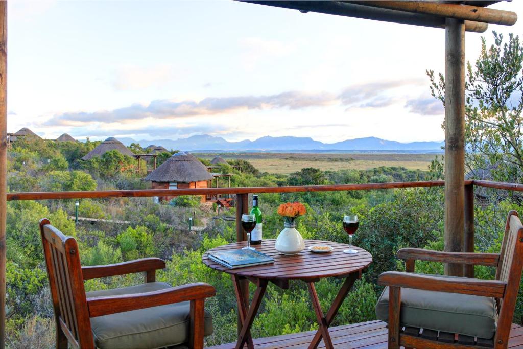 A wooden balcony with two chairs and a table set with wine, glasses, flowers, and books overlooks lush greenery, thatched-roof huts, and distant mountains—an idyllic retreat often found at Game Lodges in Western Cape.