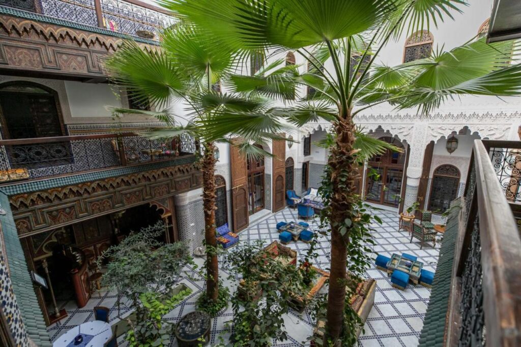 7 UNIQUE Riads in Fez You Won't Forget 28 Open courtyard of a traditional riad in Fez with palm trees, patterned tile floors, arched wooden doors, intricate mosaic walls, and colorful seating areas arranged around the garden-like space.