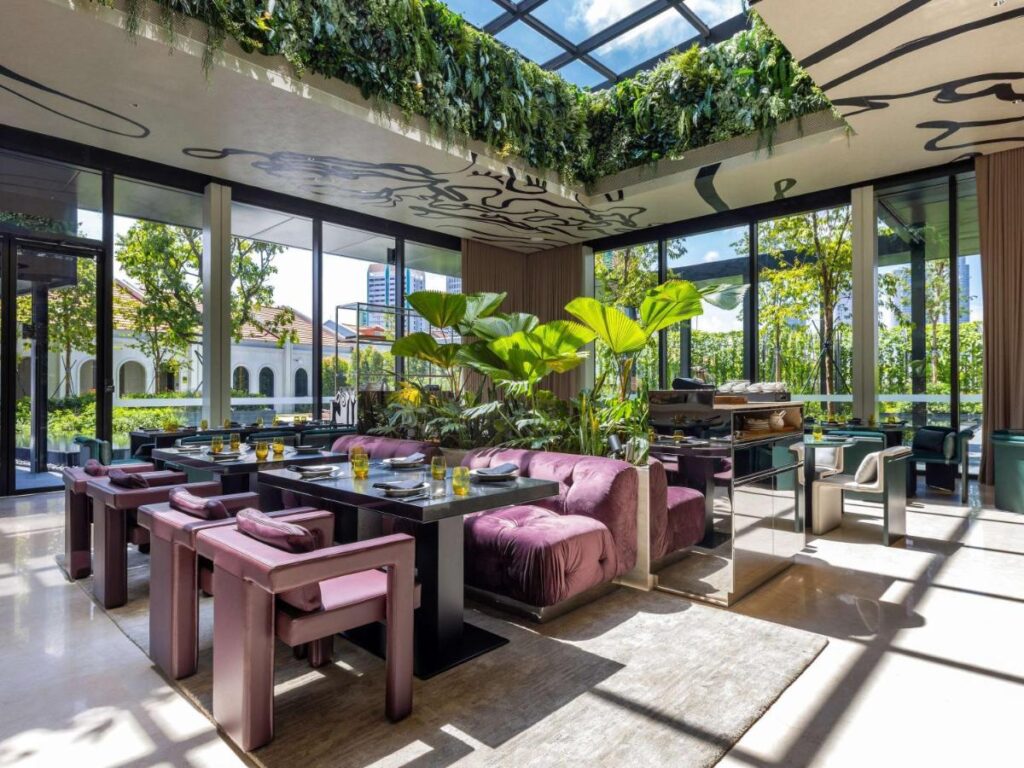 13 Most Unique Hotels in Singapore for an Incredible Holiday 49 A modern, sunlit dining area with glass walls and ceiling, purple chairs, a plush sofa, and lush greenery evokes the vibrant ambiance found in unique hotels in Singapore.