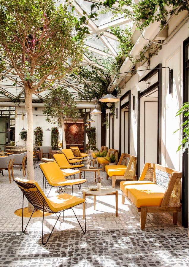 Where to Stay in Madrid: 14 Best Hotels 10 A bright, sunlit indoor patio with cobblestone flooring, lush green trees, yellow cushioned chairs, wooden benches, and marble-topped tables under a glass ceiling with hanging plants—an ideal spot for those seeking where to stay in Madrid first time.