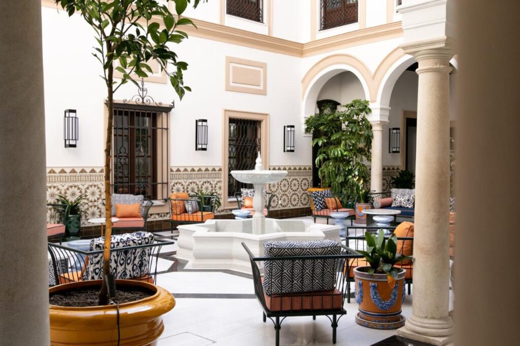 10 UNIQUE Hotels with Pool in Seville 18 A bright, elegant courtyard in the style of hotels with pool in Seville, featuring patterned tile walls, potted plants, cushioned chairs, and a white stone fountain in the center, all surrounded by graceful arches and columns.