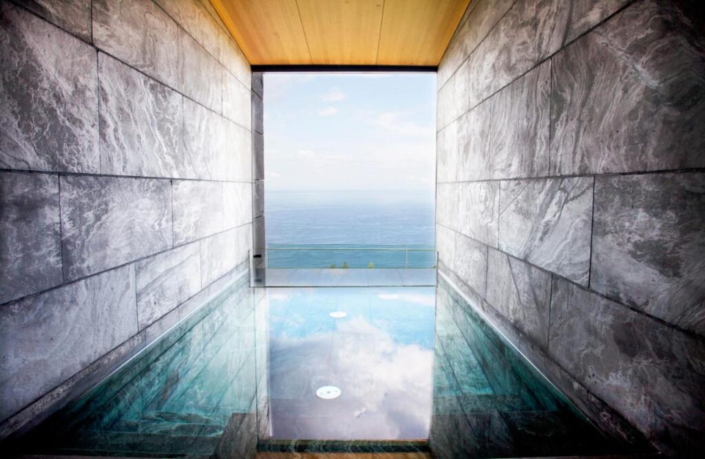 Best Boutique Hotels in San Sebastian 26 A modern indoor pool with gray marble walls and a wooden ceiling, reminiscent of the elegant designs found in boutique hotels in San Sebastian, overlooks a balcony with glass railing and a scenic view of the ocean and sky in the distance.