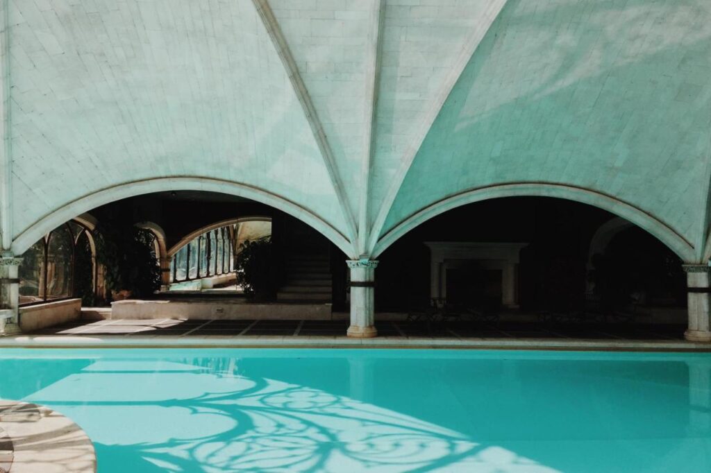25 Weekend Trips from Madrid Max. 2h Away 55 A turquoise indoor pool sits beneath a vaulted ceiling with arched pillars, perfect for relaxing on weekend trips from Madrid. Sunlight creates intricate shadows on the water and walls, with plants, a staircase, and a white fireplace in the background.