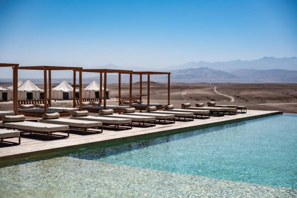 8 SPECTACULAR Agafay Desert Camps 35 A luxurious outdoor pool with lounge chairs and wooden cabanas overlooks the Agafay Desert camps, set against a stunning backdrop of distant mountains under a clear blue sky.