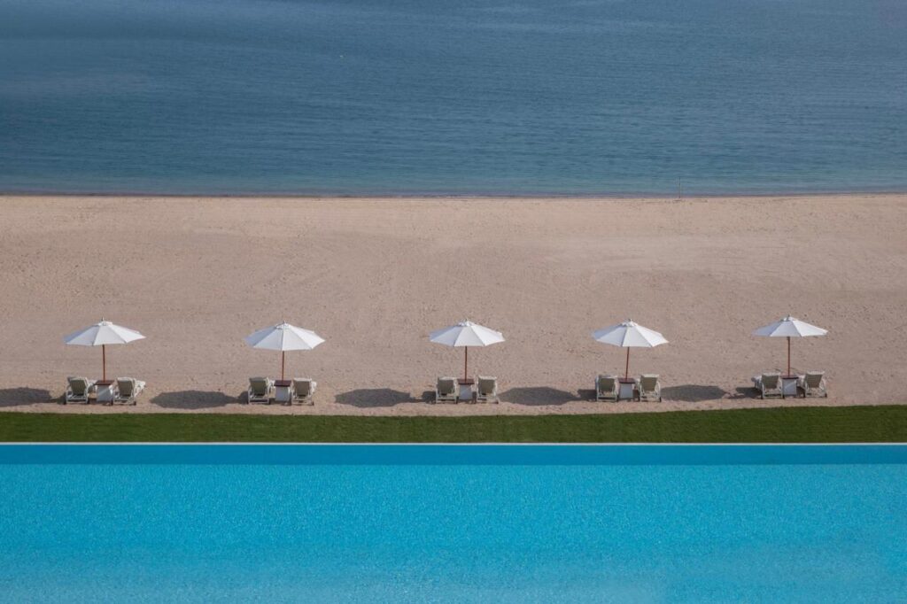 Where to Stay in Doha, Qatar: Transit, City & Beach Hotels 4 A row of white beach umbrellas and lounge chairs lines an empty sandy beach beyond a bright blue pool, with the calm sea and sky in the background—an inviting scene for anyone seeking where to stay in Doha.