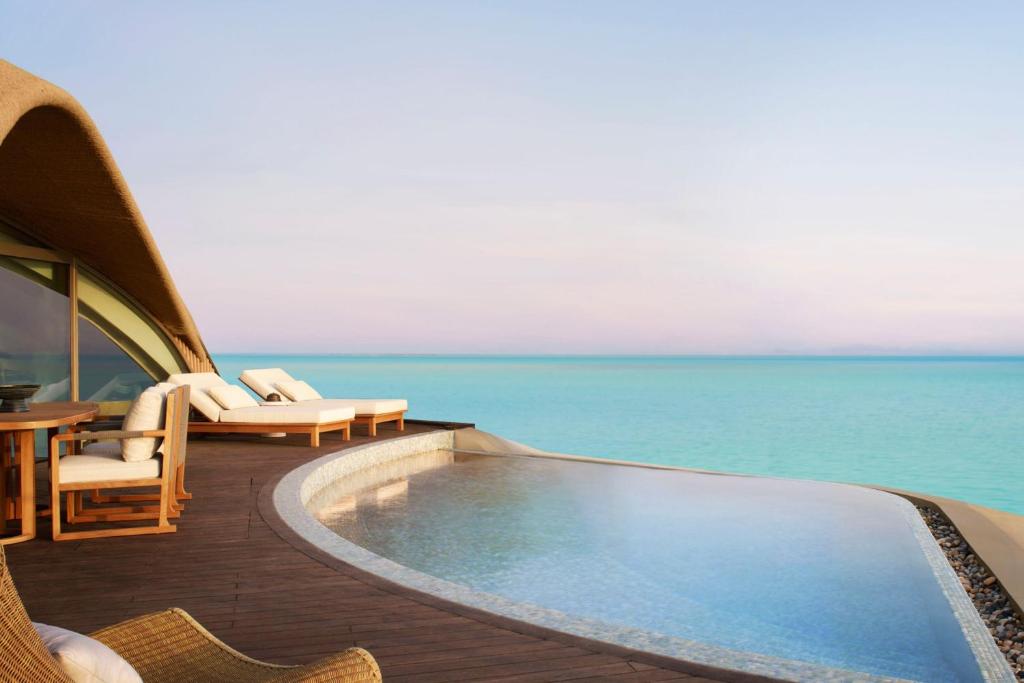 9 Best Hotels in Saudi Arabia: Unique Stays Across the Country 33 A luxurious outdoor deck at one of the Best Hotels in Saudi Arabia features lounge chairs and a curved infinity pool overlooking a calm, turquoise ocean under a pastel sky at sunset or sunrise.