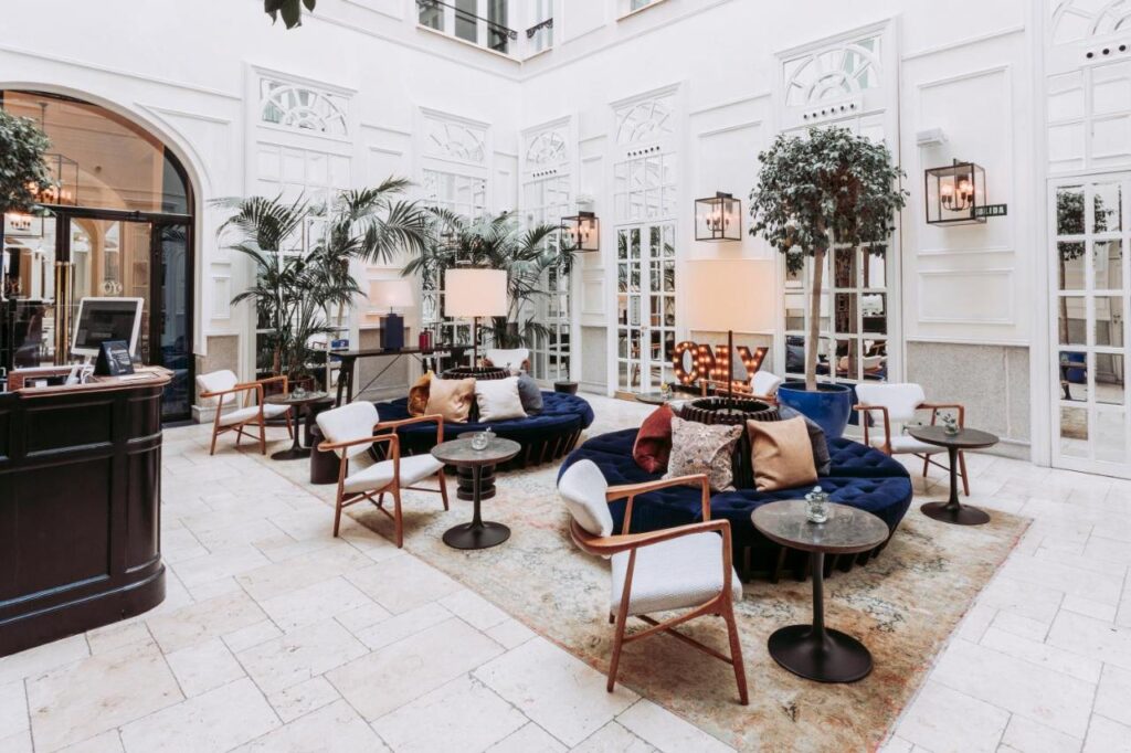 Where to Stay in Madrid: 14 Best Hotels 46 Modern, elegant lounge with plush blue sofas, white armchairs, and tall potted plants—an inviting space for those wondering where to stay in Madrid first time. Large windows and stylish decor create a bright, airy atmosphere with soft lighting throughout.