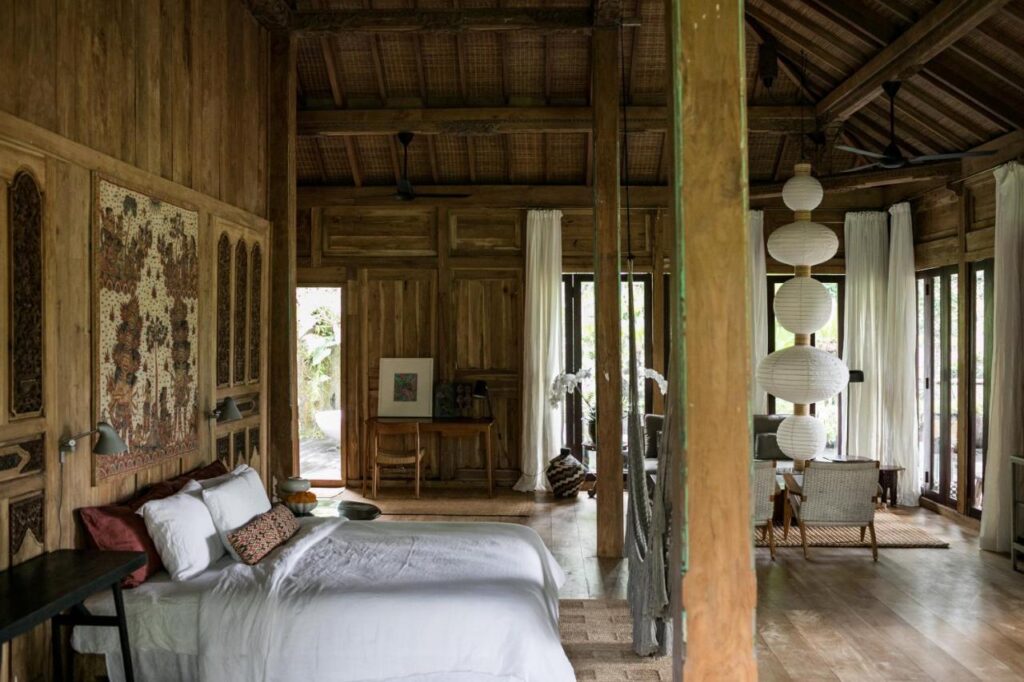 13 Most UNIQUE Places to Stay in Ubud 9 A spacious rustic bedroom with wooden walls and ceiling, a white bed, large windows with sheer curtains, a wooden desk, and hanging lanterns—one of the unique places to stay in Ubud, offering a cozy, natural atmosphere with outdoor views.