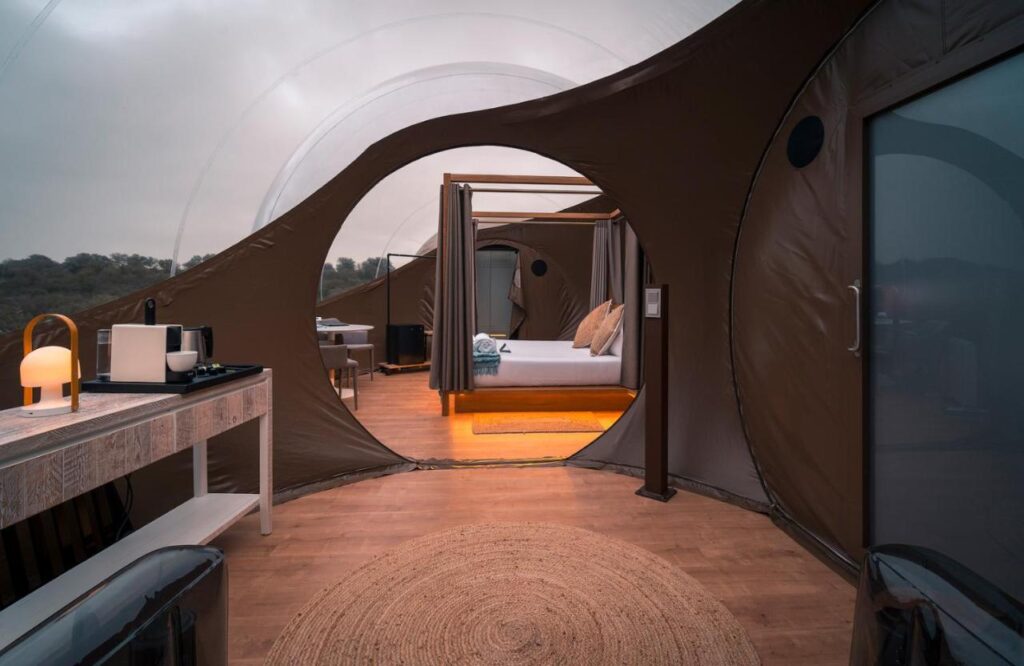 25 Weekend Trips from Madrid Max. 2h Away 21 A modern glamping dome with circular openings shows a cozy bedroom with a canopy bed, soft lighting, and a woven rug—perfect for weekend trips from Madrid. A table with a lamp, mugs, and coffee maker sits invitingly in the foreground.
