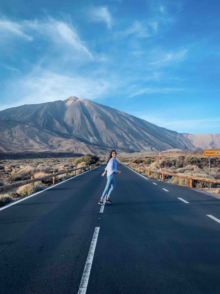 11 UNIQUE Boutique Hotels in Tenerife 2 A person in casual clothes stands joyfully in the middle of an empty road, with a large mountain and blue sky in the background—a scene reminiscent of escapes to boutique hotels in Tenerife, surrounded by dry landscape and scattered bushes.
