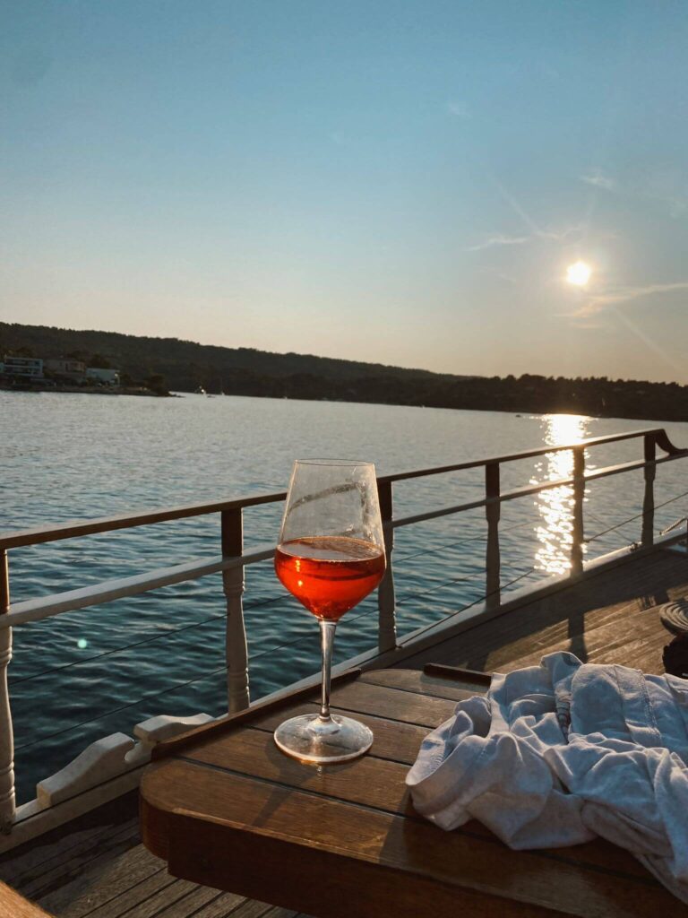 8 TOP Boutique Hotels in Split 36 A glass of rosé wine sits on a wooden table by the railing of a boat, overlooking calm water with the sun setting in the background—a perfect moment to consider where to stay in Split. A white shirt is draped over the table.
