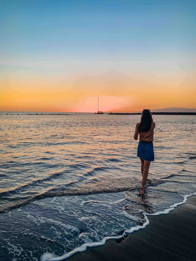11 UNIQUE Boutique Hotels in Tenerife 53 A person stands at the edge of the ocean, gazing at a colorful sunset with a sailboat in the distance—just steps from where boutique hotels in Tenerife line the shore beneath blue-to-orange skies and gentle, lapping waves.
