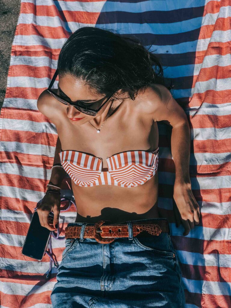 11 UNIQUE Boutique Hotels in Tenerife 49 Woman in sunglasses and a red-and-white striped bikini top lies on a matching towel, wearing blue jeans and a brown belt, holding a phone while relaxing outdoors in the sun—dreaming of Boutique Hotels in Tenerife.