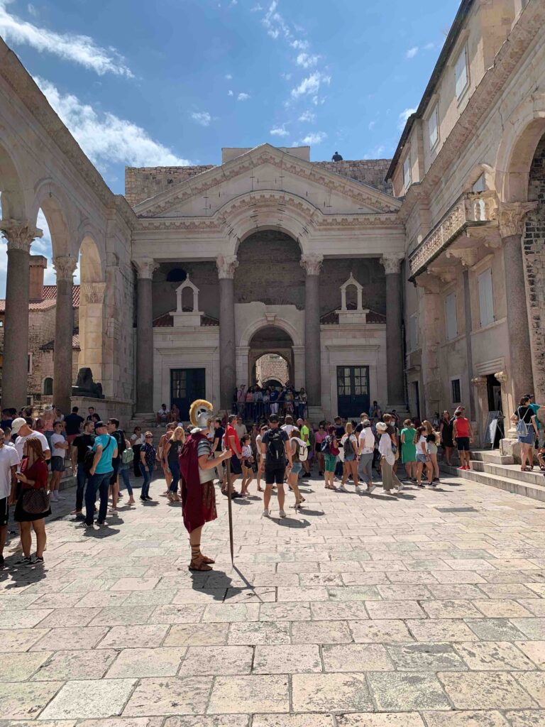 8 TOP Boutique Hotels in Split 3 A large crowd gathers in a sunny ancient courtyard with stone columns and arches. A person dressed as a Roman soldier stands in the foreground, holding a spear, while tourists exploring Where to Stay in Split admire the historic site.
