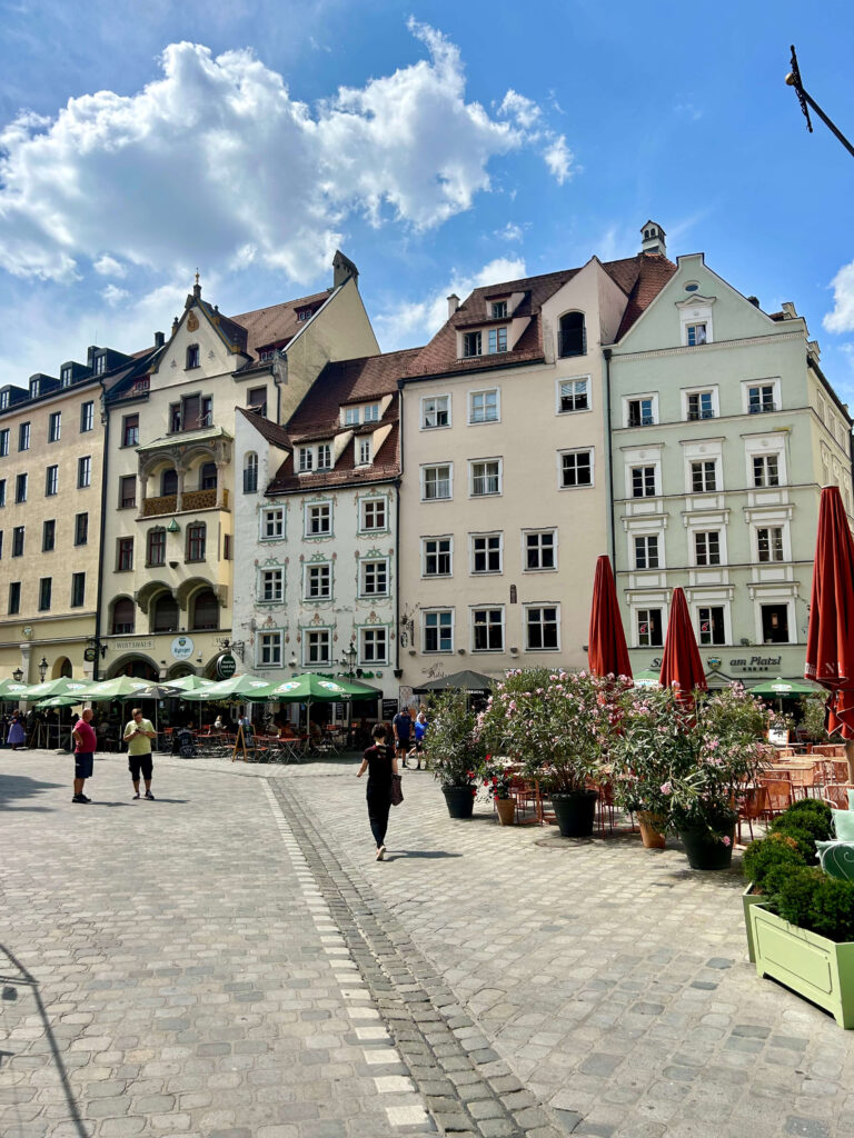 Authentic Boutique Hotels in Munich 3 A European plaza with historic buildings, boutique hotels in Munich nearby, outdoor café seating under red umbrellas, potted plants, and people walking on a sunny day with a partly cloudy sky.
