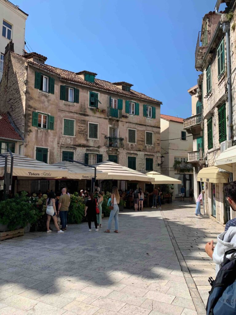 8 TOP Boutique Hotels in Split 2 People walk and gather near outdoor cafes with umbrellas in a sunlit European square, surrounded by old stone buildings with green shutters—a charming scene for anyone wondering Where to Stay in Split under a clear blue sky.