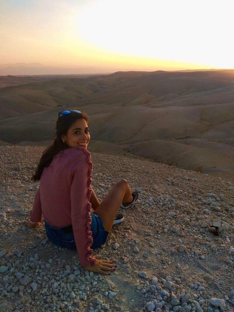 8 SPECTACULAR Agafay Desert Camps 8 A woman sits on a rocky hillside at sunset, looking back and smiling. She wears a pink sweater and shorts, with sunglasses on her head. The scene recalls the tranquil beauty of Agafay Desert camps under a golden sky.