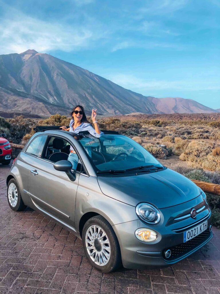 11 UNIQUE Boutique Hotels in Tenerife 3 A smiling woman wearing sunglasses stands through the sunroof of a gray Fiat 500 parked on a scenic road, reminiscent of adventures near boutique hotels in Tenerife, with mountains and dry brush in the background under a clear blue sky.