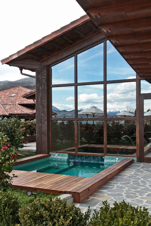 A small outdoor pool with wooden decking sits next to a modern house, reminiscent of boutique hotels in Asturias, its large glass windows reflecting mountains, sky, nearby umbrellas, and lush greenery.