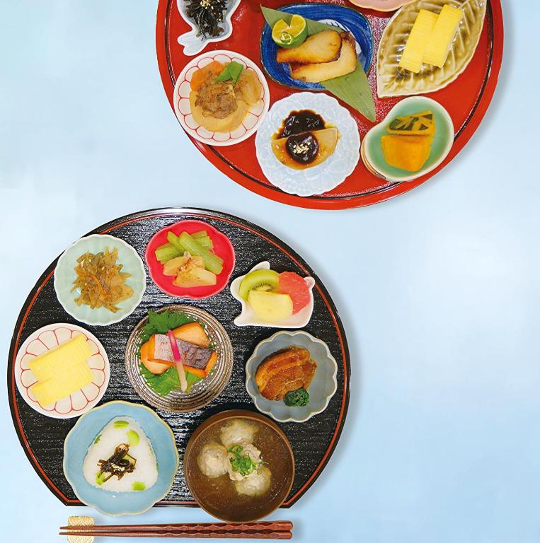 8 BEST Ryokans in Osaka 18 Two round trays with assorted Japanese dishes, like those served at ryokans in Osaka, feature sliced fruits, grilled fish, vegetables, tamagoyaki, soup, pickles, and rice in small colorful bowls. A pair of chopsticks lies beside the black tray.