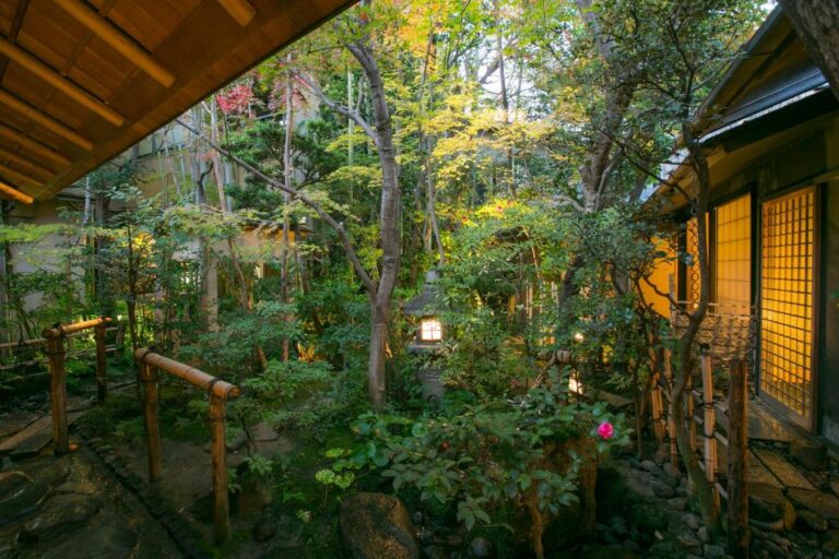 A serene Japanese garden with lush green trees, bamboo fences, stone lanterns, and warm lights glowing from traditional wooden buildings on either side—reminiscent of the peaceful ryokans in Gion Kyoto.