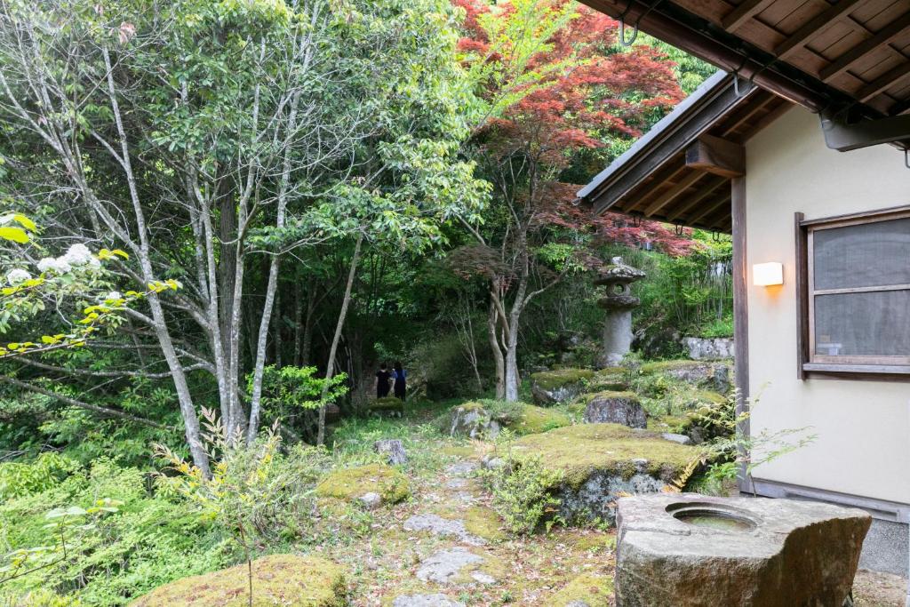 8 BEST Ryokans in Osaka 23 A lush Japanese garden with trees, moss-covered rocks, a stone lantern, and a stone basin sits beside a traditional wooden house—much like those found at ryokans in Osaka—with two people visible on the shaded pathway.