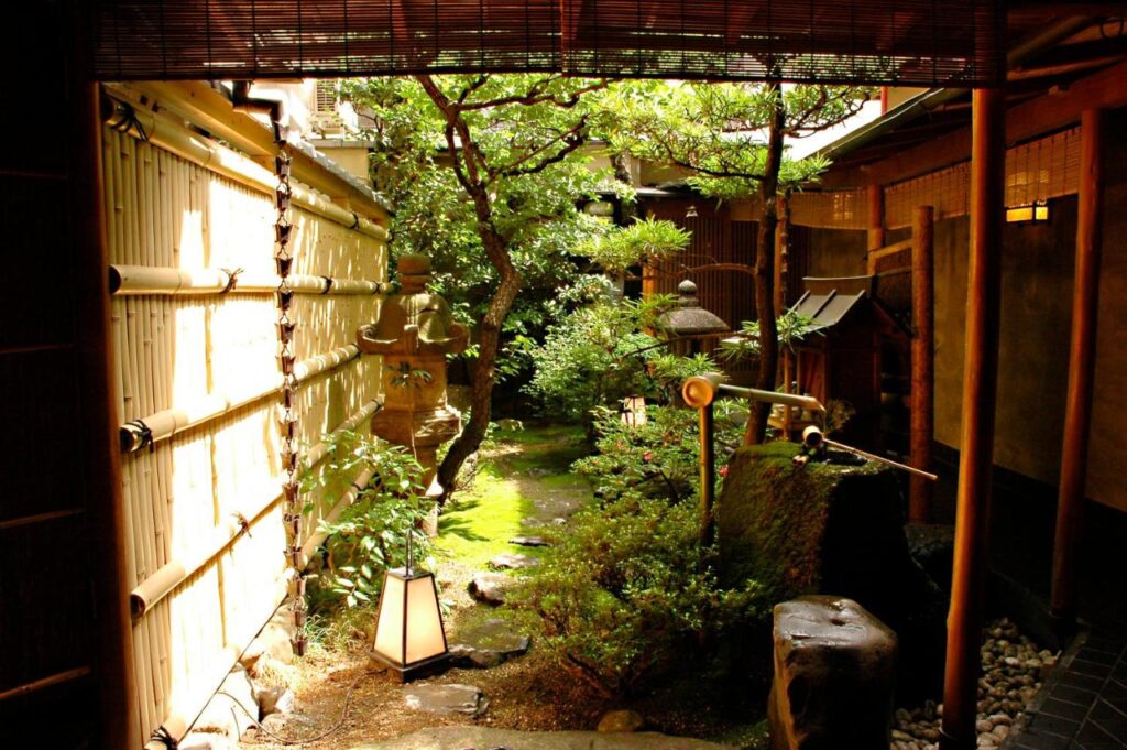 7 UNIQUE Ryokans in Gion Kyoto 10 A traditional Japanese garden with bamboo fencing, stone lanterns, lush green plants, and a bamboo water feature evokes the peaceful atmosphere found at ryokans in Gion Kyoto, where soft sunlight filters through tranquil surroundings.