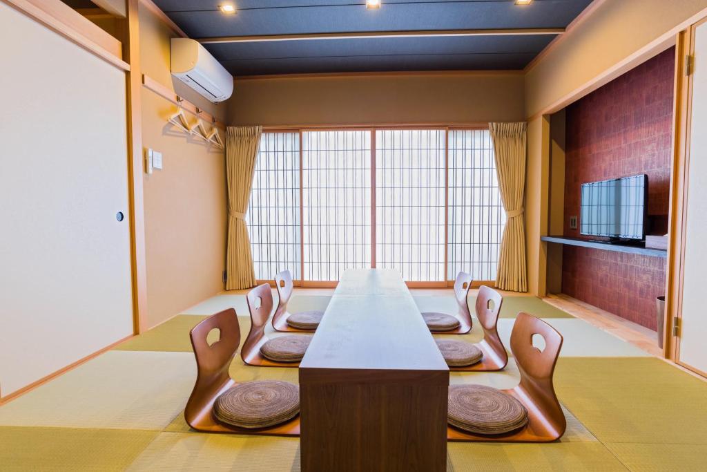 8 BEST Ryokans in Osaka 16 A traditional Japanese-style room inspired by ryokans in Osaka, featuring tatami mats, a low wooden table, six floor chairs with cushions, sliding shoji doors, natural light from large windows, and a wall-mounted TV in an alcove.
