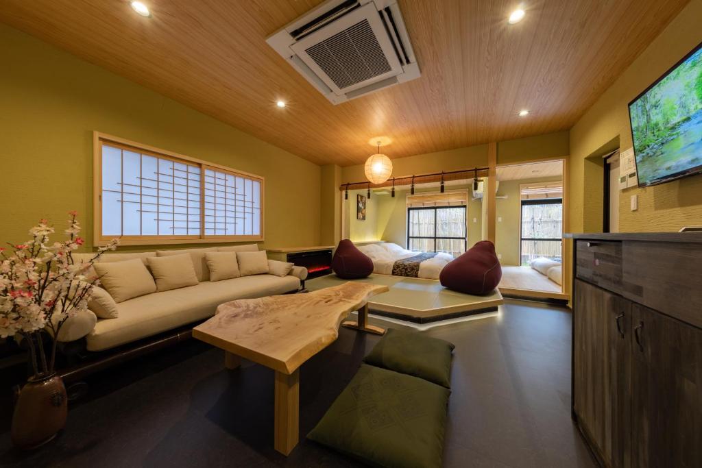 8 BEST Ryokans in Osaka 6 A cozy Japanese-style room inspired by ryokans in Osaka, featuring a low wooden table, floor cushions, a beige sofa, cherry blossom decor, raised tatami area with beds, and soft lighting; a TV is mounted on the wall.
