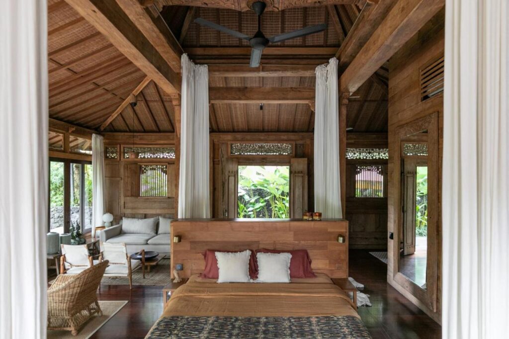 13 Most UNIQUE Places to Stay in Ubud 34 A cozy bedroom with a wooden interior, high ceiling, large windows, and a ceiling fan. Perfect for those seeking unique places to stay in Ubud, it offers a comfy bed, inviting sofa and chairs, and views of lush greenery outside.
