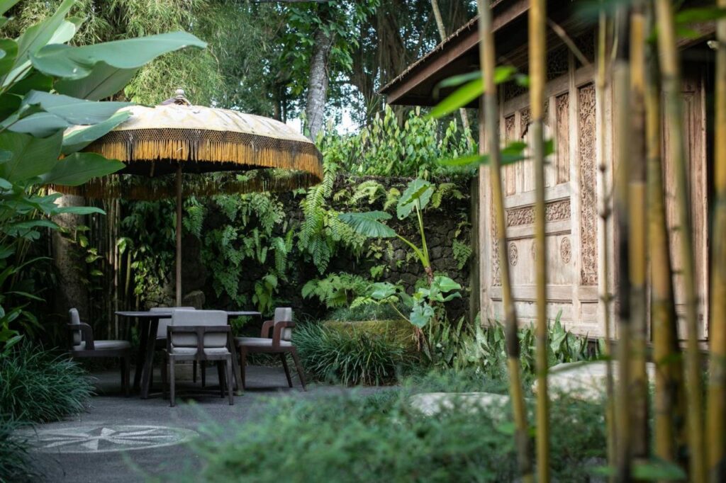 13 Most UNIQUE Places to Stay in Ubud 35 A serene garden patio with a round table and four chairs under a large decorative umbrella, surrounded by lush greenery and bamboo, offers one of the most unique places to stay in Ubud beside a wooden building with intricate carvings.