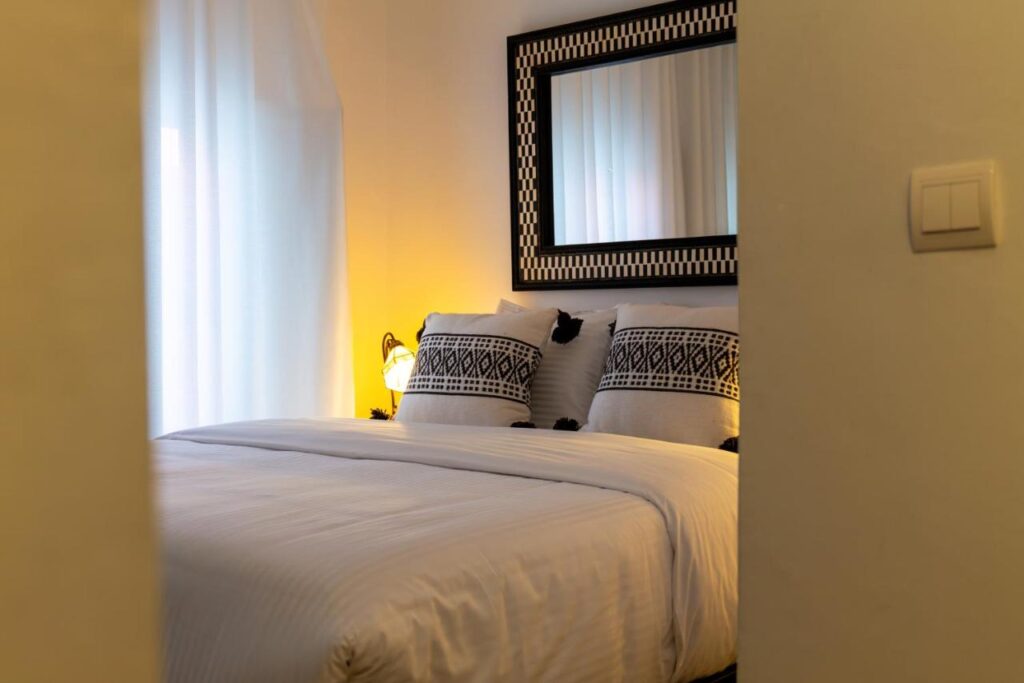 9 UNIQUE Riads in Tangier 21 A cozy bedroom inspired by riads in Tangier, featuring a neatly made bed, white bedding, black-and-white patterned pillows, a checkered mirror above the bed, soft lighting, and sheer curtains at the window.