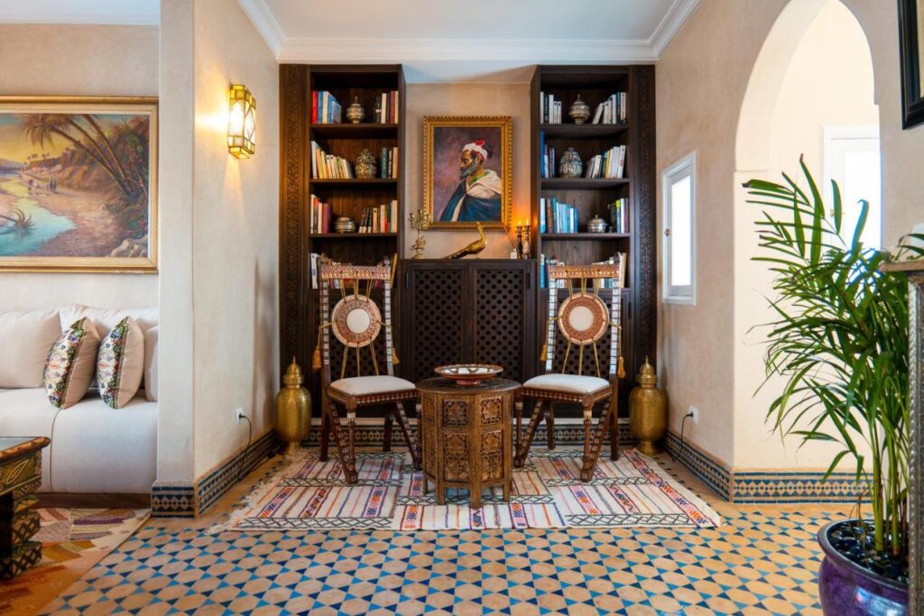 9 UNIQUE Riads in Tangier 19 A cozy, Moroccan-style sitting area inspired by riads in Tangier, with two ornate wooden chairs, a small round table, bookshelves, a framed portrait, patterned rugs, tiled floors, golden vases, and a decorative plant on the right.
