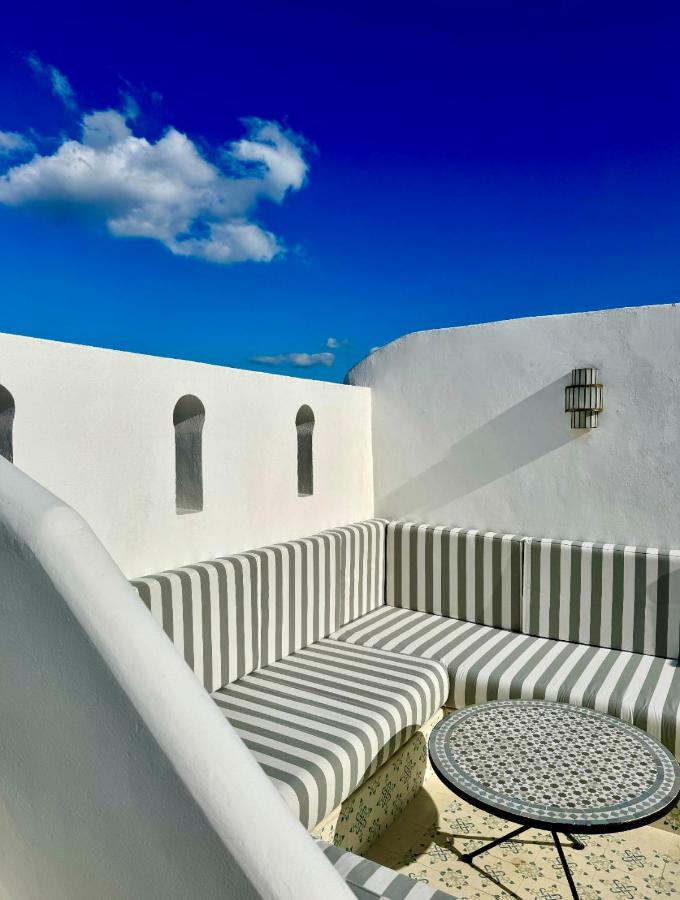 9 UNIQUE Riads in Tangier 20 A sunlit outdoor patio inspired by riads in Tangier, with white walls, a striped cushioned bench, a round mosaic table, and a deep blue sky with a single fluffy cloud above.