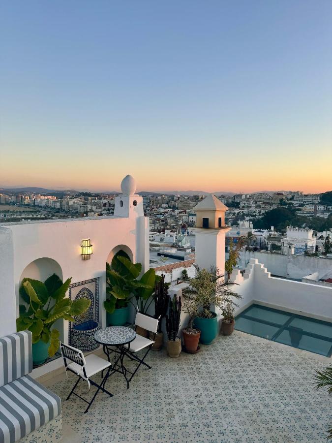 9 UNIQUE Riads in Tangier 18 A rooftop terrace inspired by riads in Tangier, with patterned tiles, potted plants, a small table with chairs, and city buildings in the background under a clear sky at sunset.