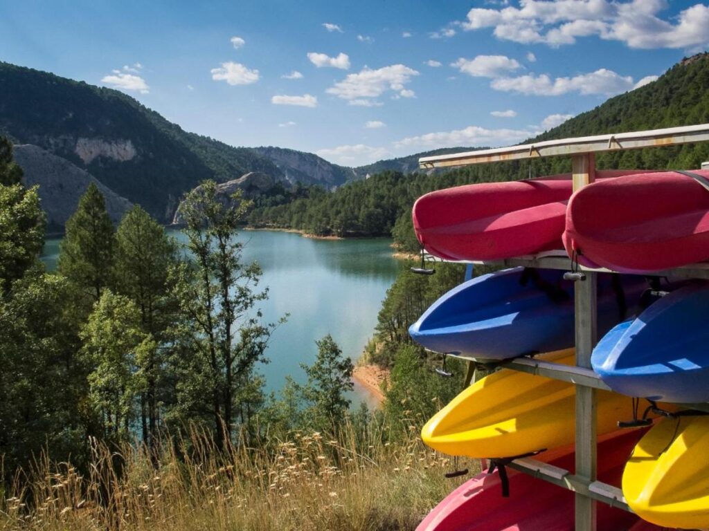 25 Weekend Trips from Madrid Max. 2h Away 84 Colorful kayaks are stacked on a rack by a calm lake surrounded by green trees and mountains under a partly cloudy sky. This peaceful outdoor scene is perfect inspiration for weekend trips from Madrid. Tall grass and pine trees frame the view.