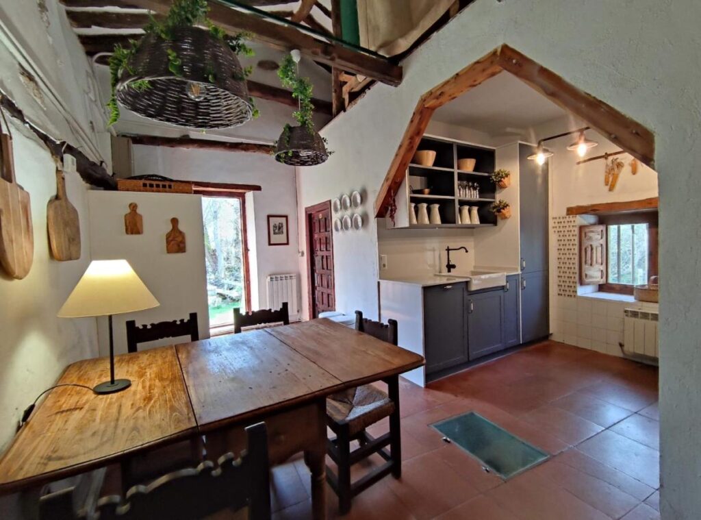 25 Weekend Trips from Madrid Max. 2h Away 68 A rustic kitchen and dining area, perfect for planning weekend trips from Madrid, features wooden beams, a wooden table with chairs, hanging wicker lights, cutting boards on the wall, open shelves with dishes, and ample natural light from a window and door.