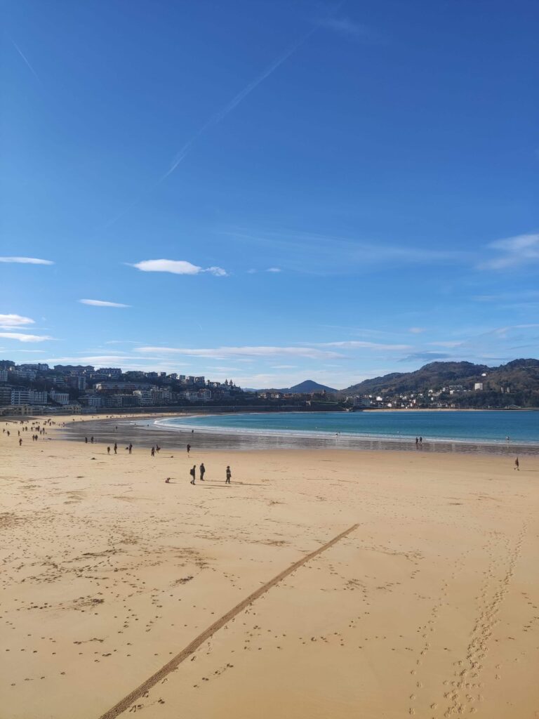 Best Boutique Hotels in San Sebastian 2 Wide sandy beach with scattered people walking, calm blue sea, and distant hills dotted with boutique hotels in San Sebastian beneath a clear blue sky with a few clouds.