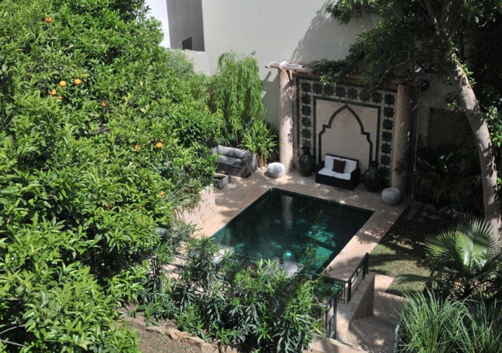 9 UNIQUE Riads in Tangier 12 A small, rectangular courtyard pool surrounded by lush green plants, orange trees, and a stone patio with modern outdoor seating against a decorative wall with an arched design, reminiscent of the tranquil riads in Tangier.