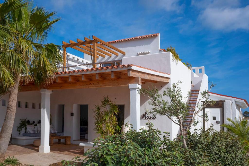 A white Mediterranean-style villa with terracotta roof tiles, wooden pergola, and shaded terrace, this serene retreat evokes the charm of boutique hotels in Formentera, surrounded by palm trees and greenery under a blue sky.