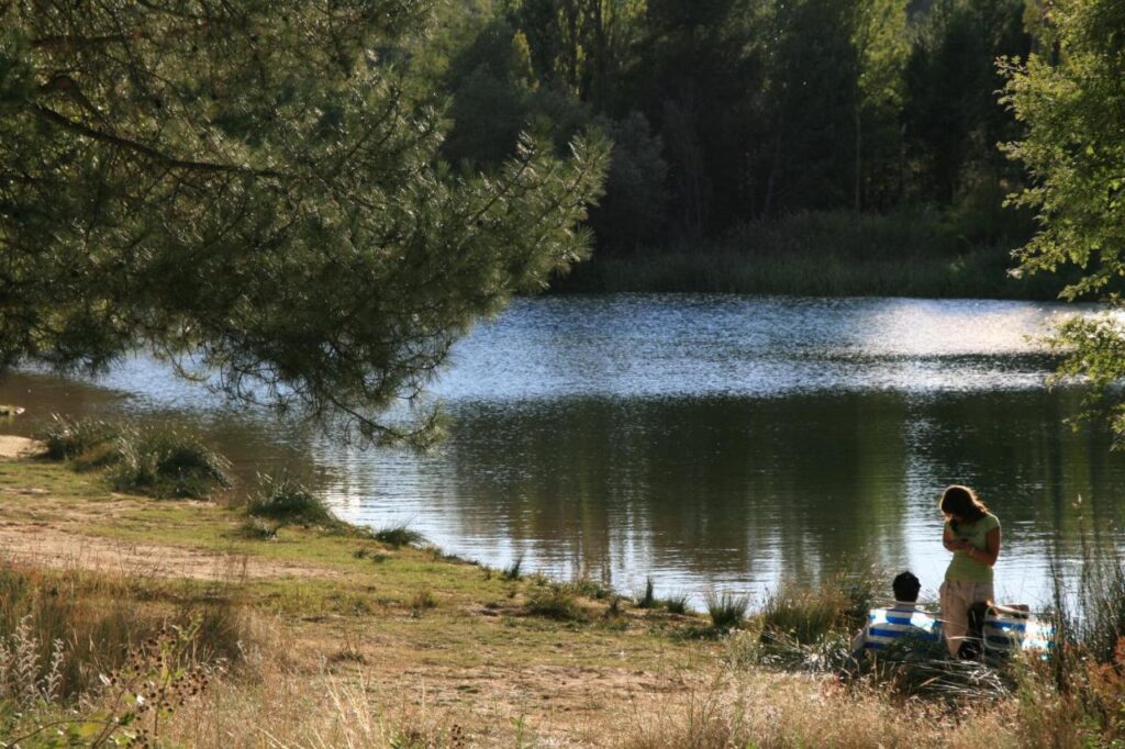 25 Weekend Trips from Madrid Max. 2h Away 83 A woman and a child stand near the edge of a calm lake surrounded by trees and grassy banks, with sunlight filtering through the branches—perfect for peaceful weekend trips from Madrid.