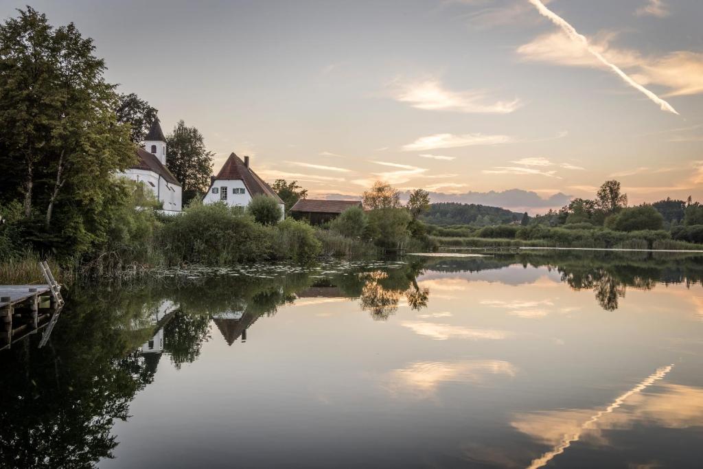 FAIRYTALE Castle Hotels in Bavaria 21 A tranquil lake at sunset reflects nearby trees, bushes, and a few white houses with steep roofs—reminiscent of the charming views found near castle hotels in Bavaria. The sky is streaked with clouds and contrails, creating a serene and peaceful atmosphere.